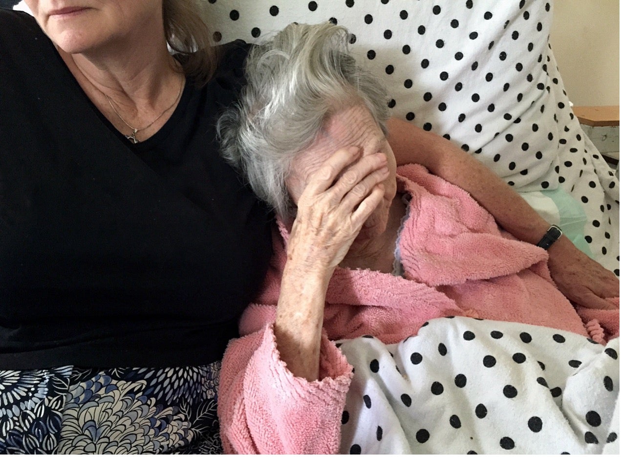 An elderly woman in a pink robe sits on a bed, covering her face with her hand. Beside her, another woman sits close, offering support. The bed is covered with a polka-dot blanket.