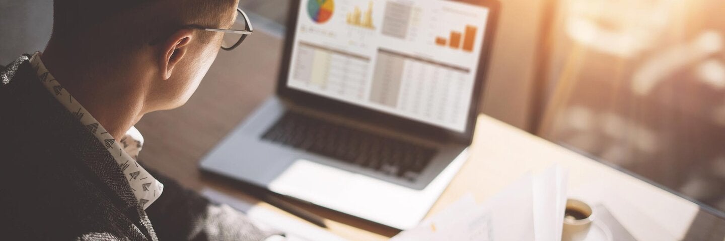 Young finance market analyst in eyeglasses working at sunny office on laptop while sitting at wooden table.Businessman analyze document in his hands.Graphs and diagramm on notebook screen.Blurred.