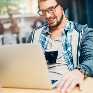 man in a beanie and glasses sitting at his laptop with a cup of coffee